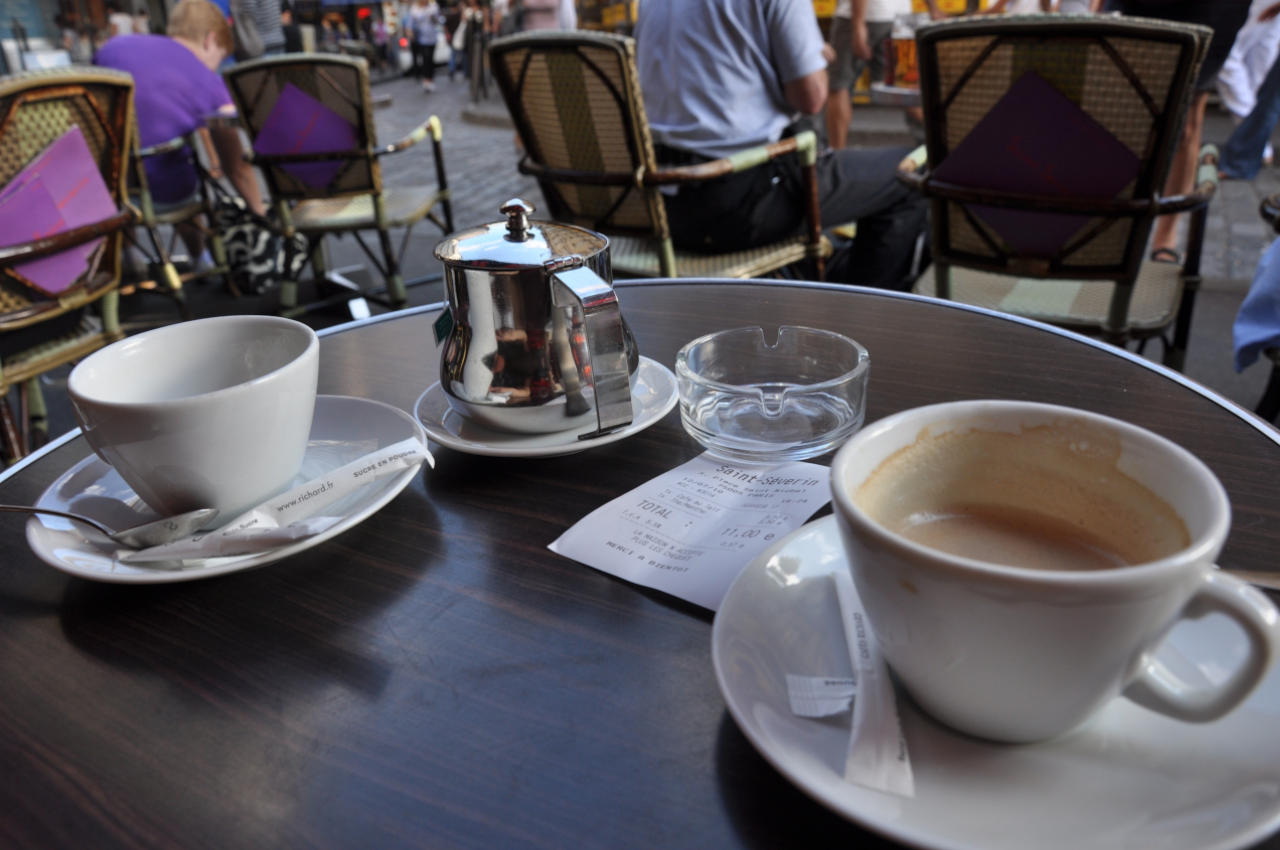 Au premier plan de la photo se trouve une table de bar avec deux tasses de café dans leur coupelle.